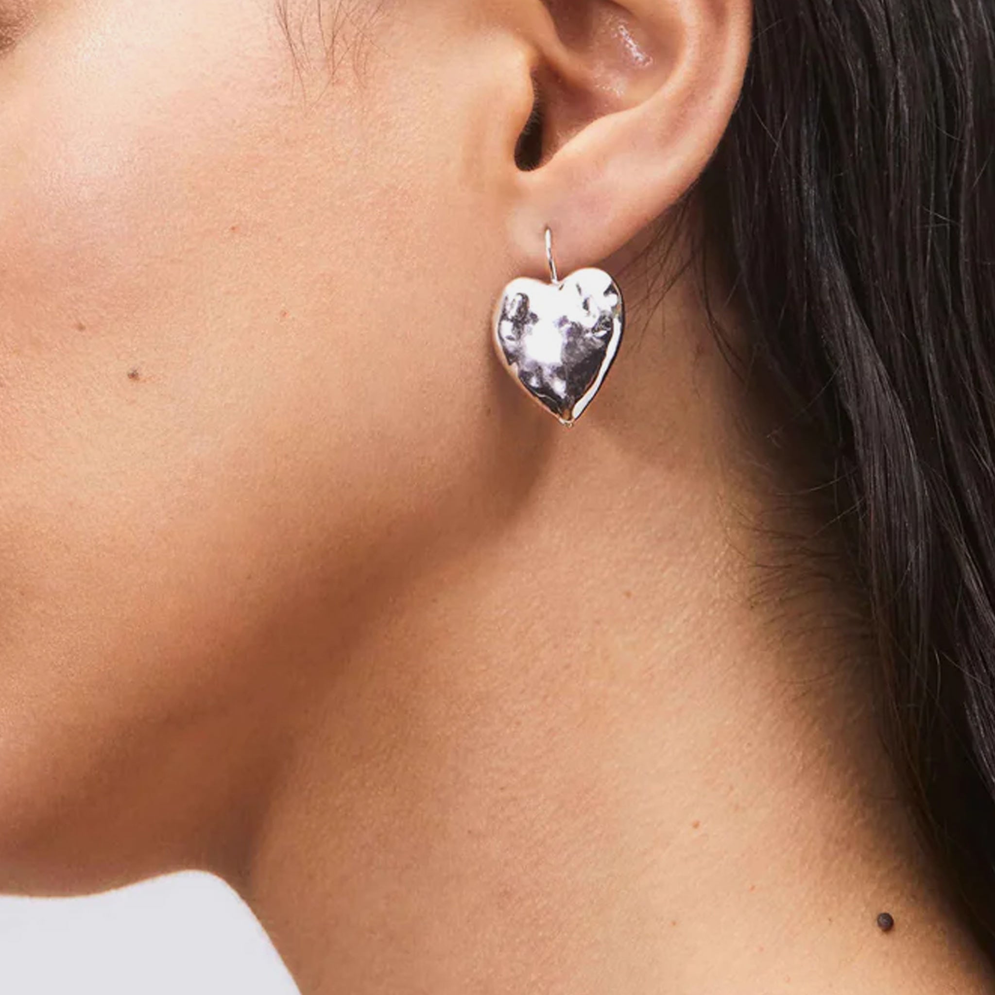 A model wears the Hear Burn Earrings - a silver hammered drop earrings in a heart shape.