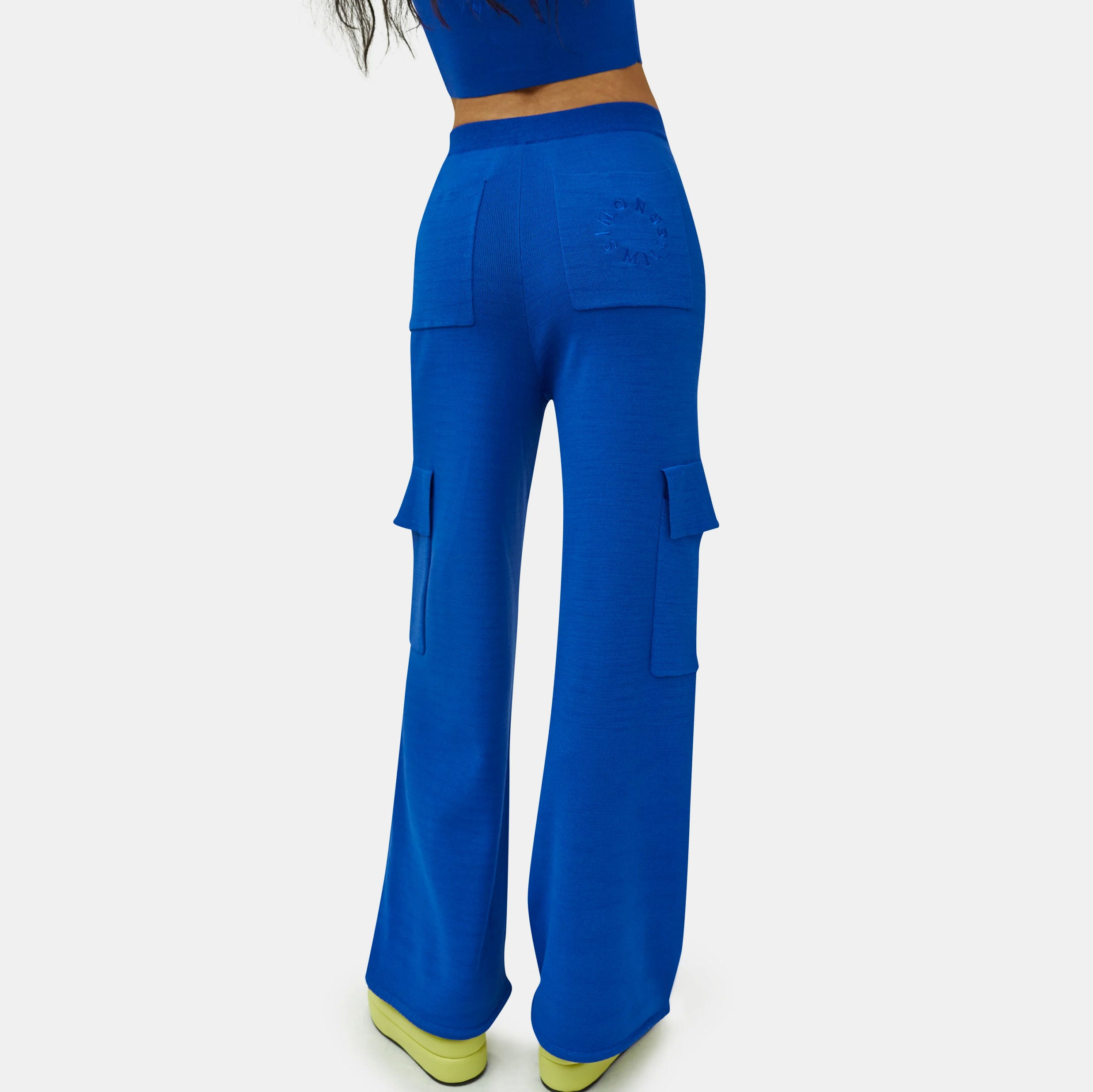 Close detail half body photo of the model wearing the Hesby Pant - a vibrant blue knit wide leg pant with side cargo-style pockets and a button & zip closure - back view.