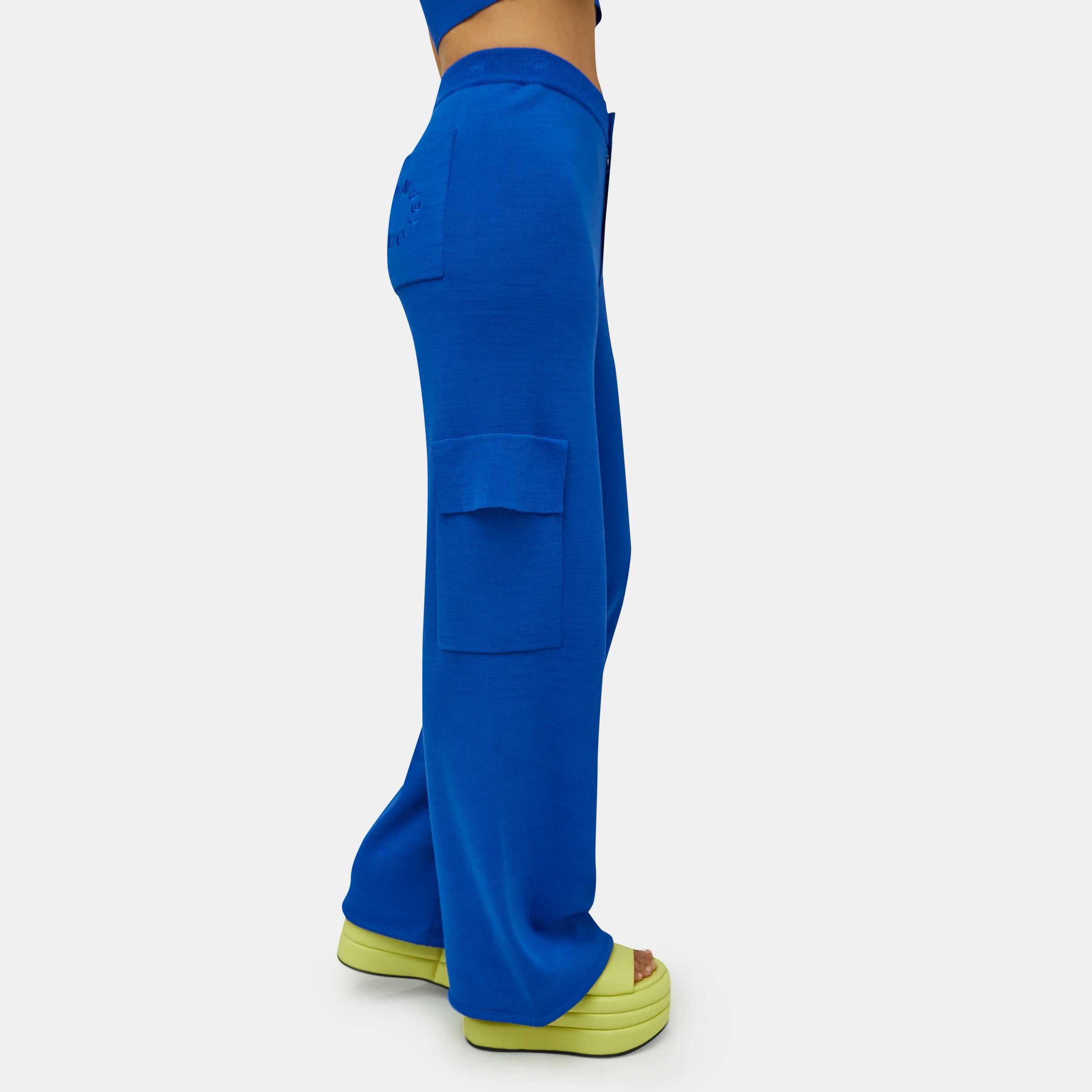 Close detail half body photo of the model wearing the Hesby Pant - a vibrant blue knit wide leg pant with side cargo-style pockets and a button & zip closure - side view.