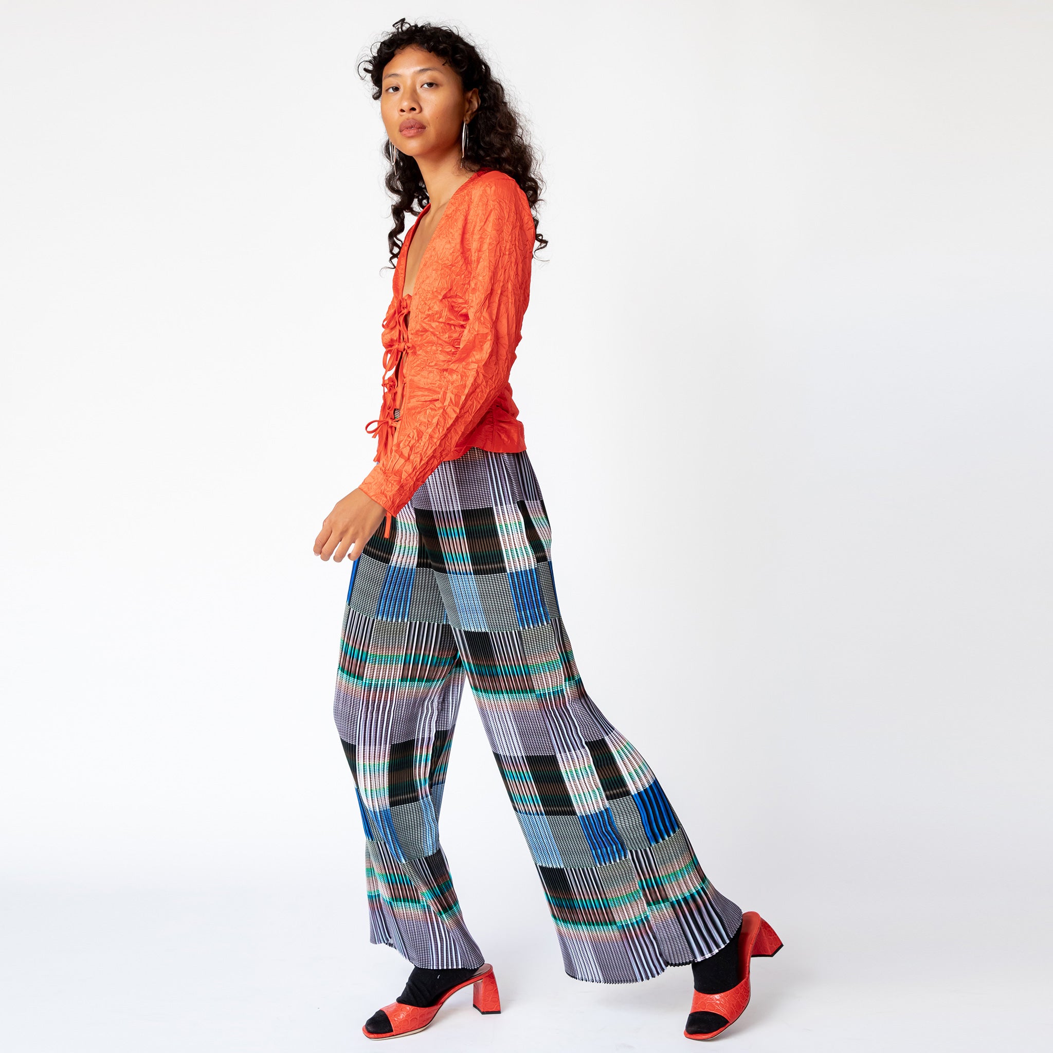 A model walks sideways while wearing the Julie Heuer pleated Jack Trousers in a gridded print pattern and a vibrant red blouse.