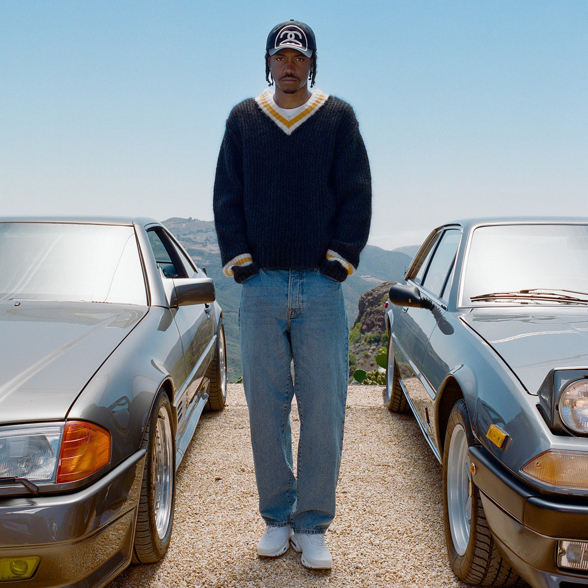 Full body photo of model standing between two cars wearing  a oversized black v neck sweater with a white and yellow line around the neck and wrists.