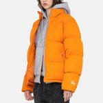A model wears the Stussy Nylon Down Puffer jacket in orange over a grey hoodie while standing at an angle.