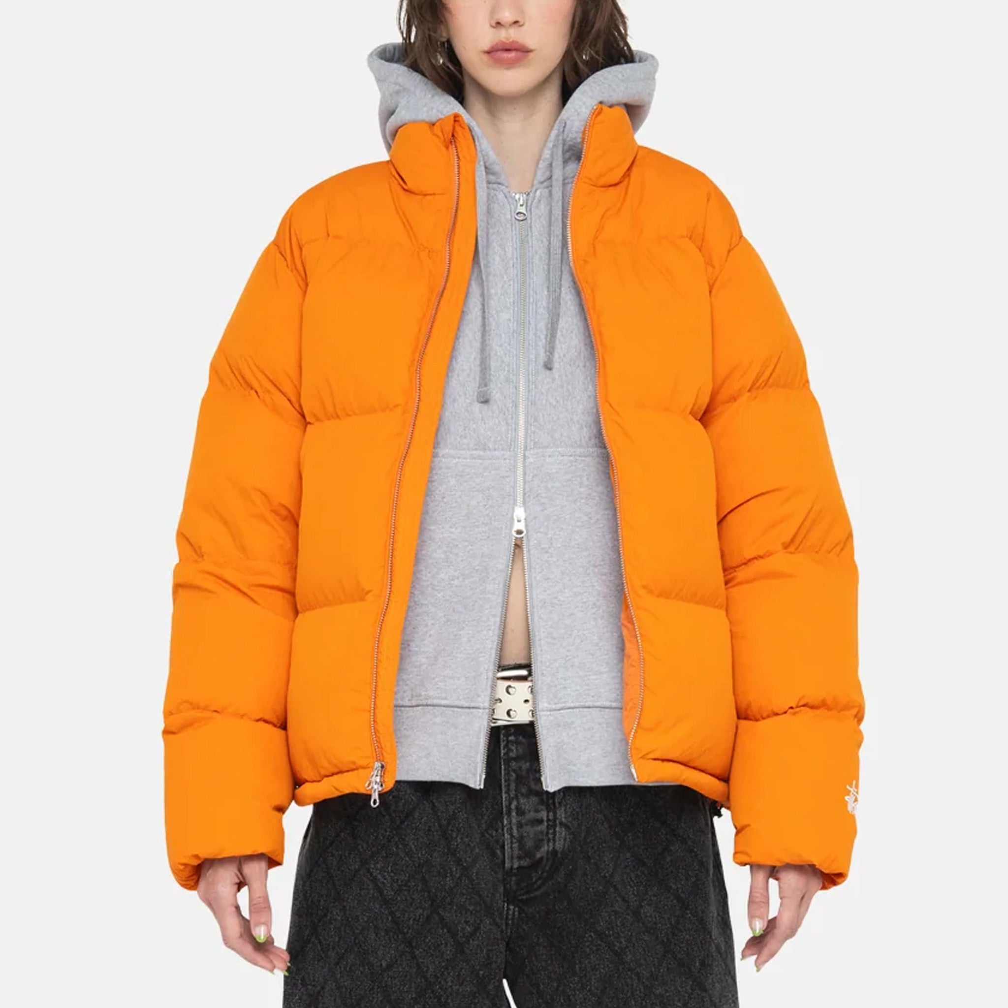 A model wears the Stussy Nylon Down Puffer jacket in orange over a grey hoodie while facing forward.