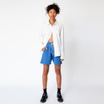 Full outfit view of the Oversized Button Down Shirt in white, worn half unbuttoned and paired with blue sweat shorts and black loafers.
