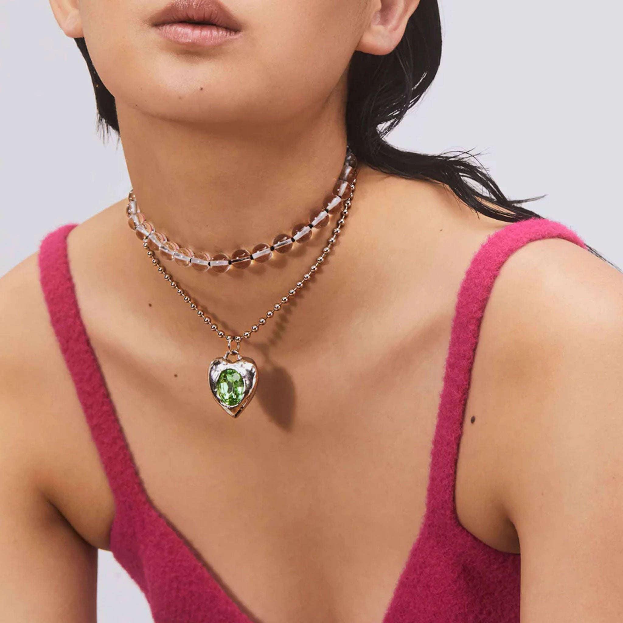 A model wears the Pacha Necklace - a silver-toned roughly shaped heart pendant necklace featuring a clear green stone.