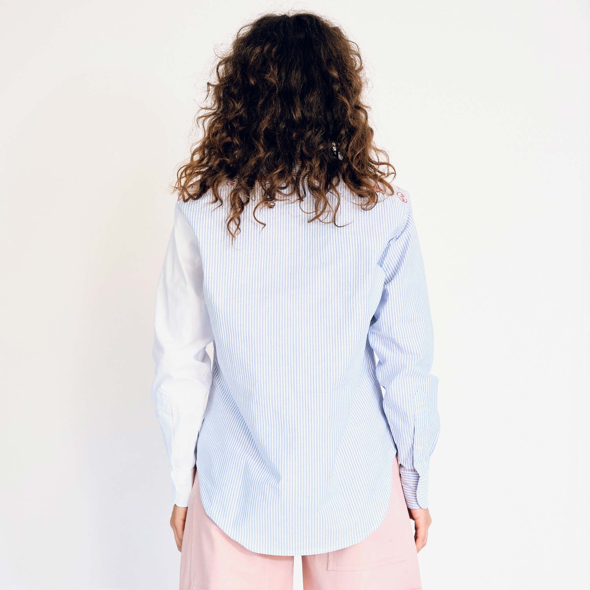 A model wears the upcycled patchwork button up shirt from Aries, which has various Aries iconography printed on the right half of the shirt and is pinstriped on the left side - back view.