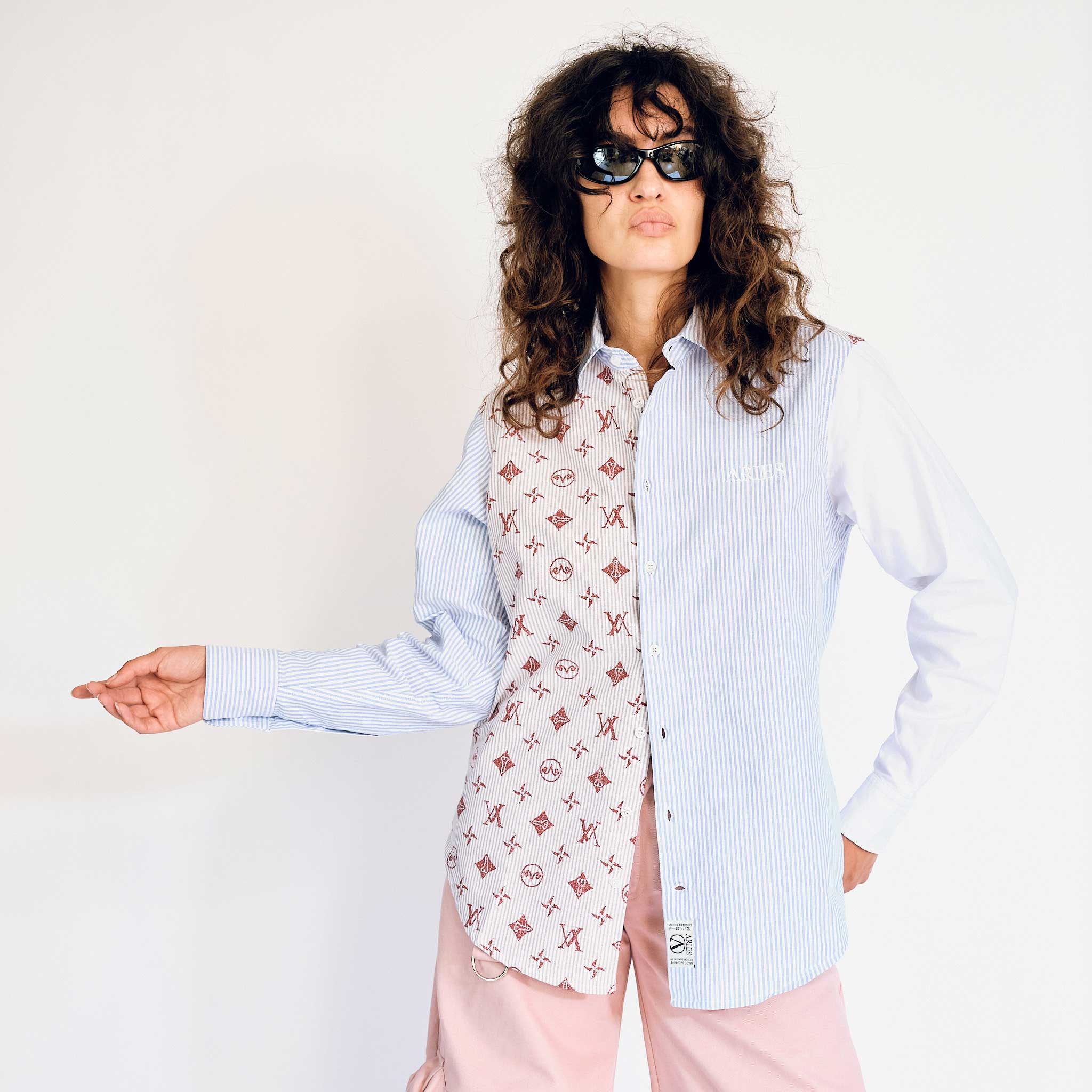A model wears the upcycled patchwork button up shirt from Aries, which has various Aries iconography printed on the right half of the shirt and is pinstriped on the left side - front view.