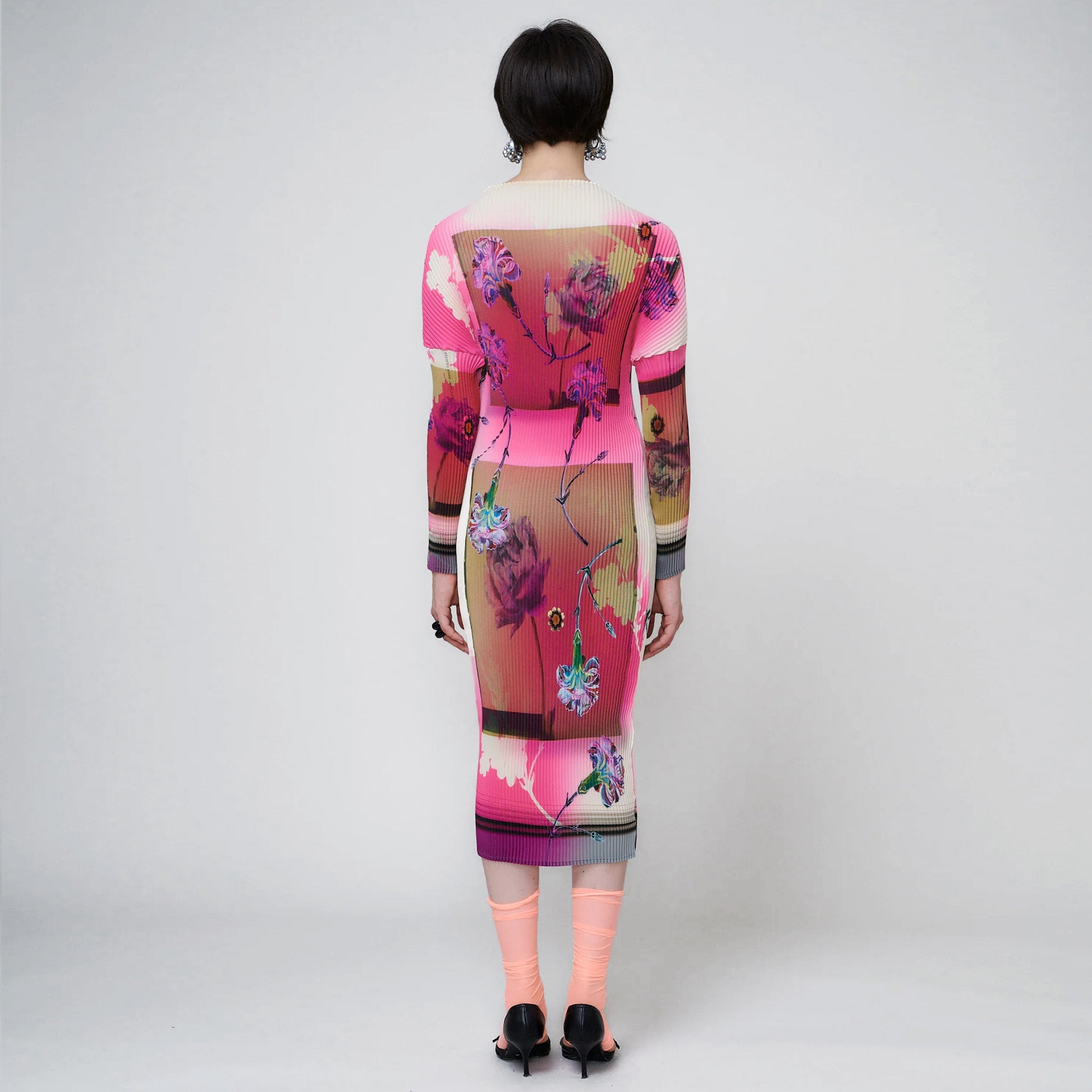 A model wears the midi-length pleated Pauki Dress in a vibrant pink and white floral print - back view.