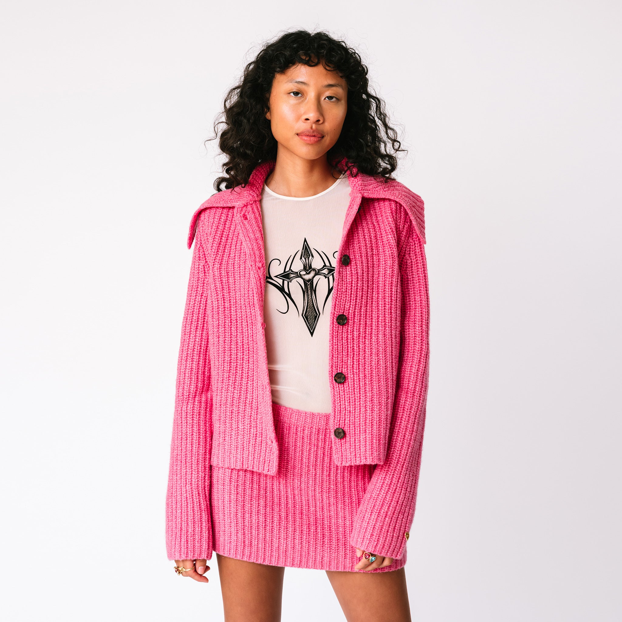 A model wears the chunky ribbed pink Paw Cardigan unbuttoned with the matching Barb ribbed skirt, front view.