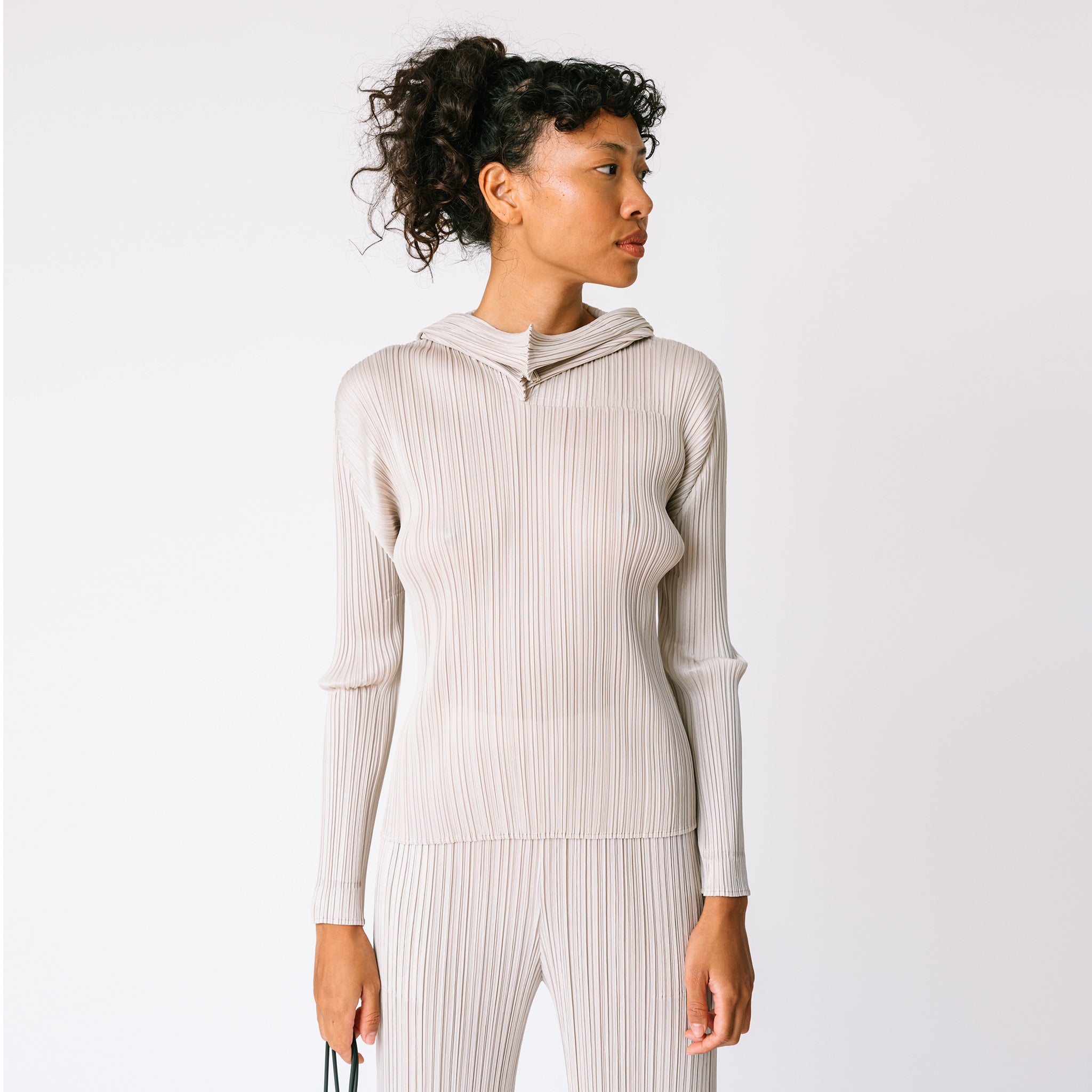 A model looks to the side while wearing the light grey Pleats Please pleated hoodie in greige, with matching pleated pants.