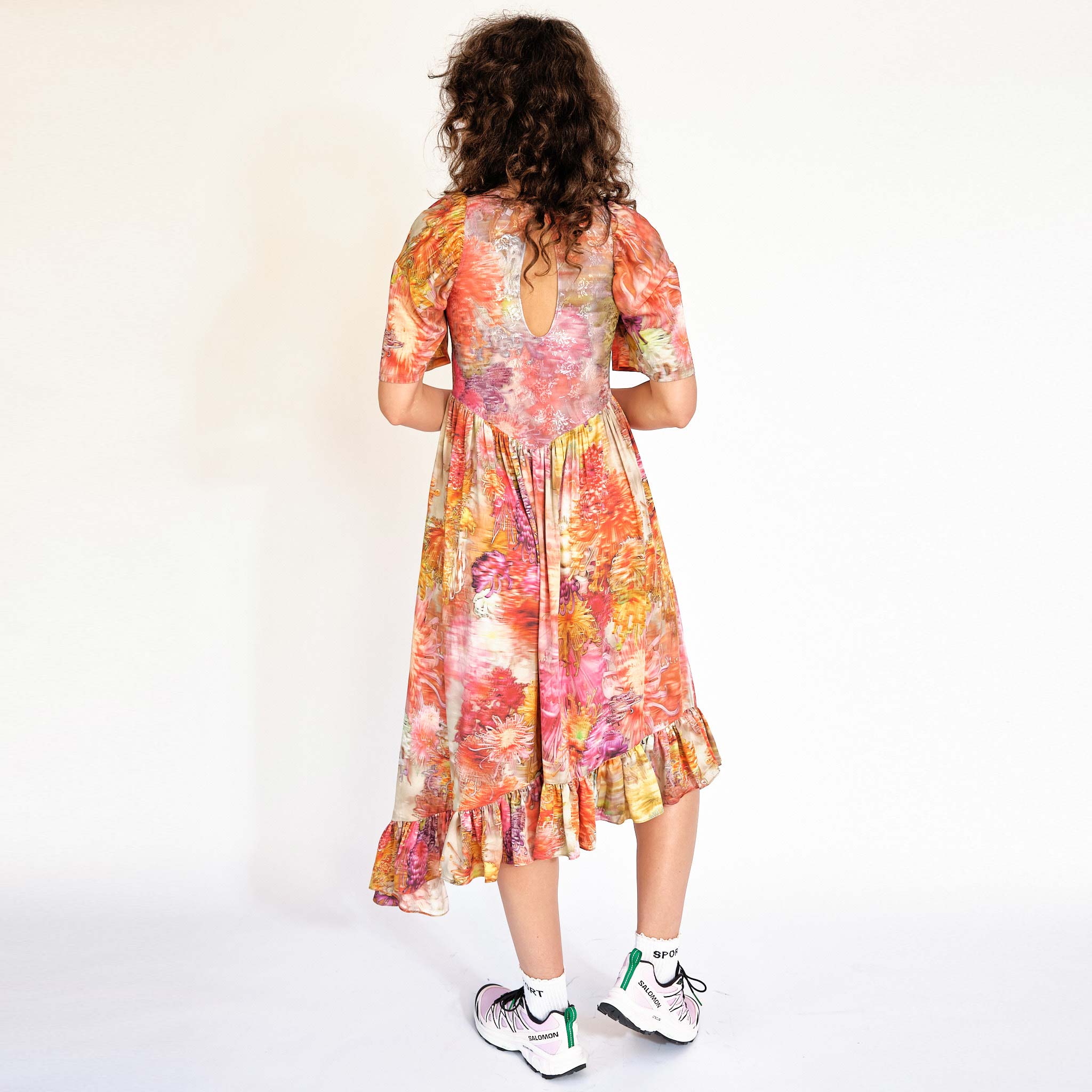 A model wears the Pollinate Dress in Chrysanthemum - multicolored floral dress with short bell sleeves and an asymmetrical ruffled hem - back view.