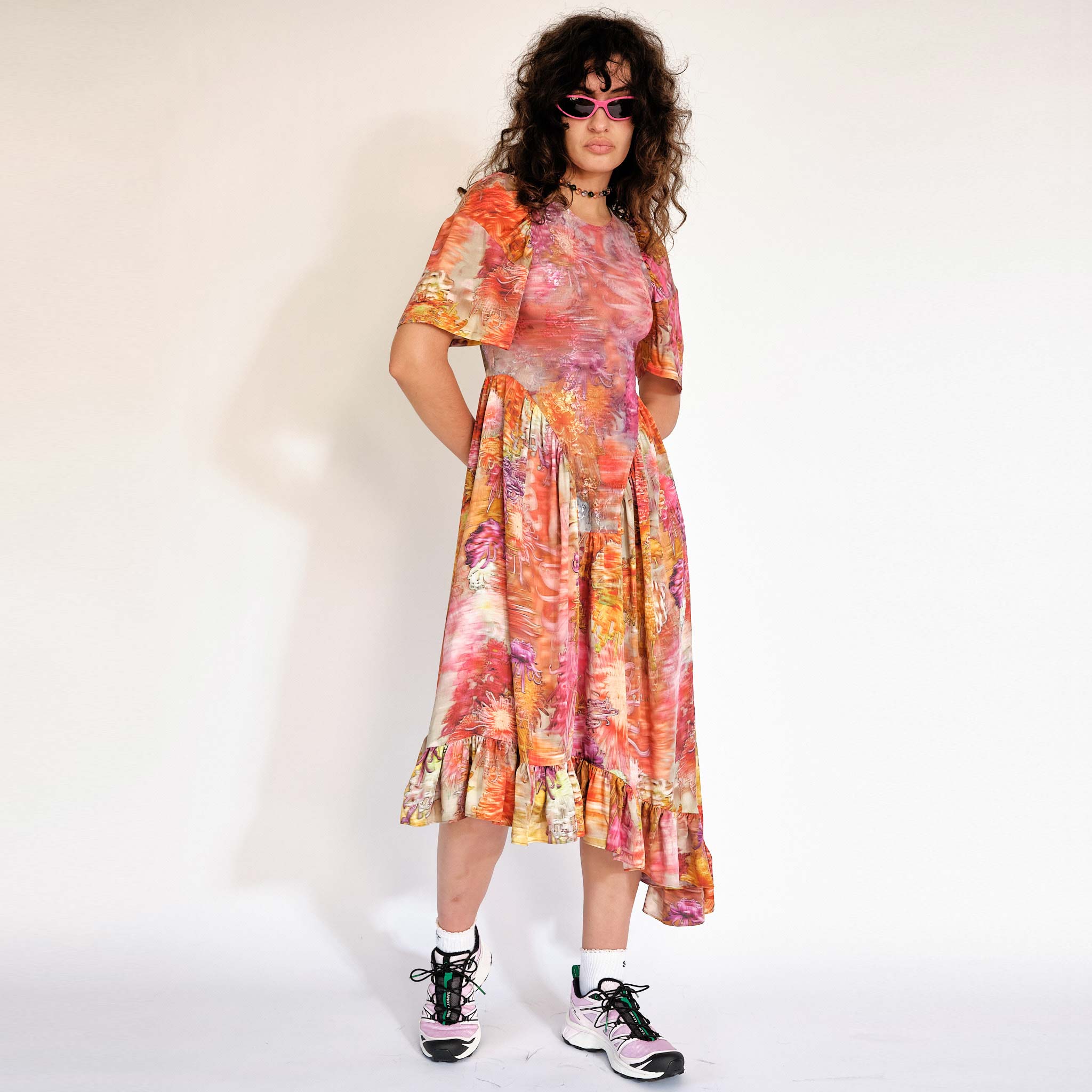 A model wears the Pollinate Dress in Chrysanthemum - multicolored floral dress with short bell sleeves and an asymmetrical ruffled hem - front view.