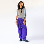 A model wears the frost gray sleeveless polo top with contrasting navy blue collar and gold button hardware, paired with vibrant purple corduroy pants.