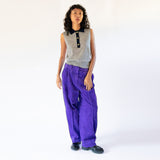 A model wears the frost gray sleeveless polo top with contrasting navy blue collar and gold button hardware, paired with vibrant purple corduroy pants.