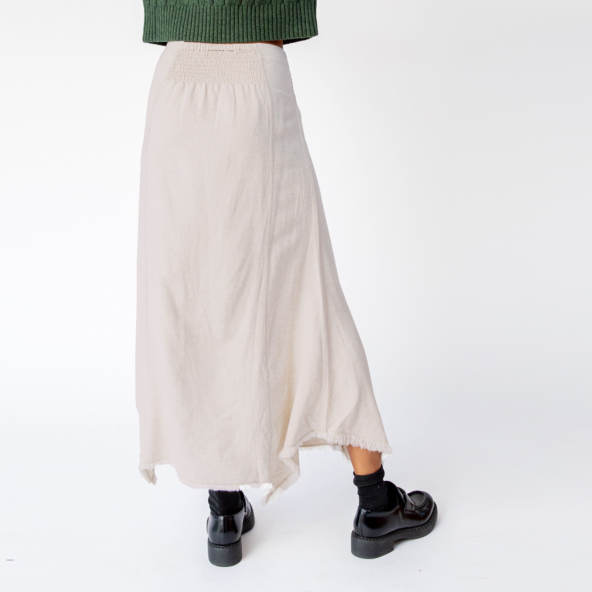 A model wears Misc Etc's smocked linen midi skirt in sand, featuring a smocked elastic waistband with drawstring and an asymmetrical hem, paired here with a green cable knit sweater and black loafers - back view.