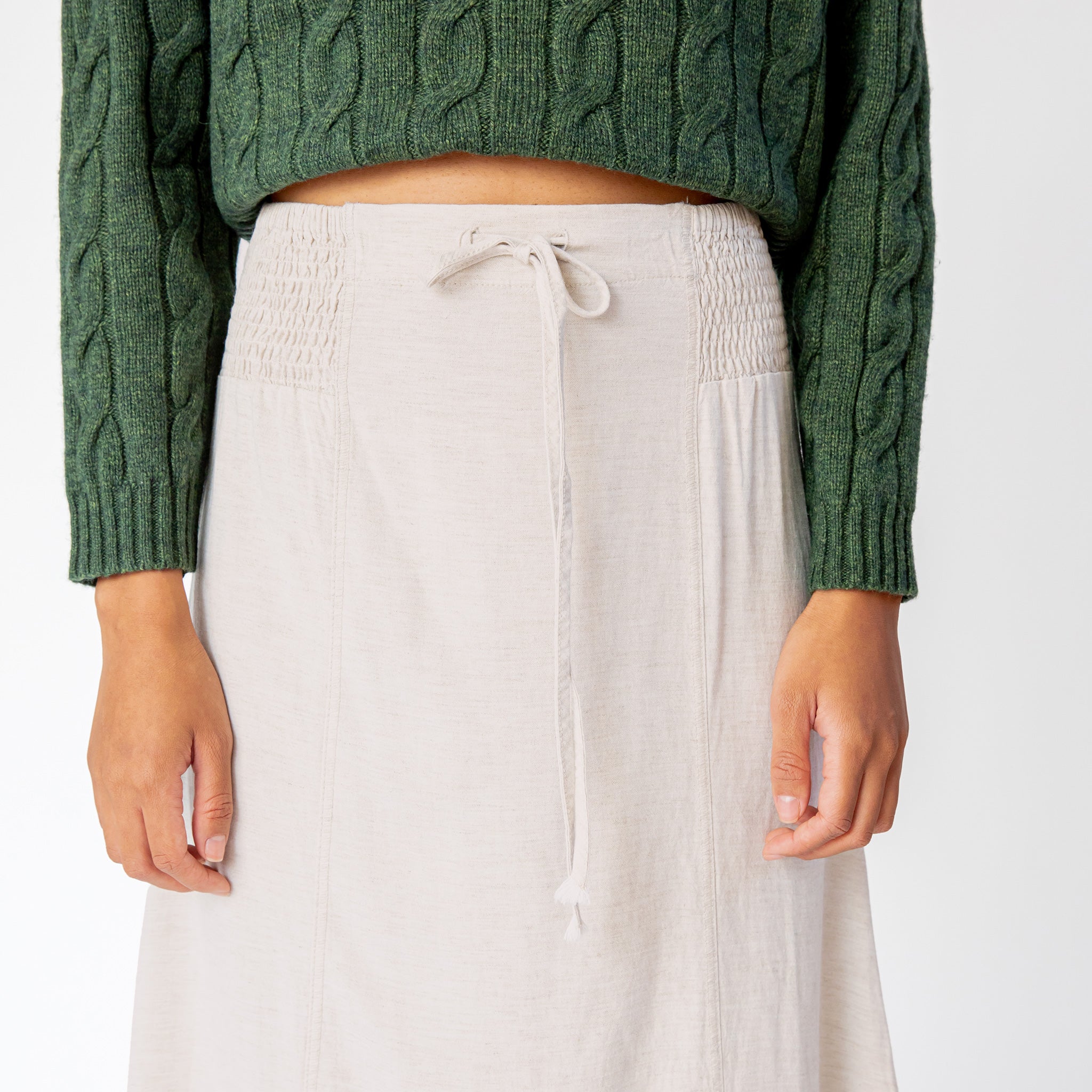 A detailed close up of the Misc Etc smocked linen midi skirt in sand, featuring a smocked elastic waistband with drawstring, paired here with a green cable knit sweater.