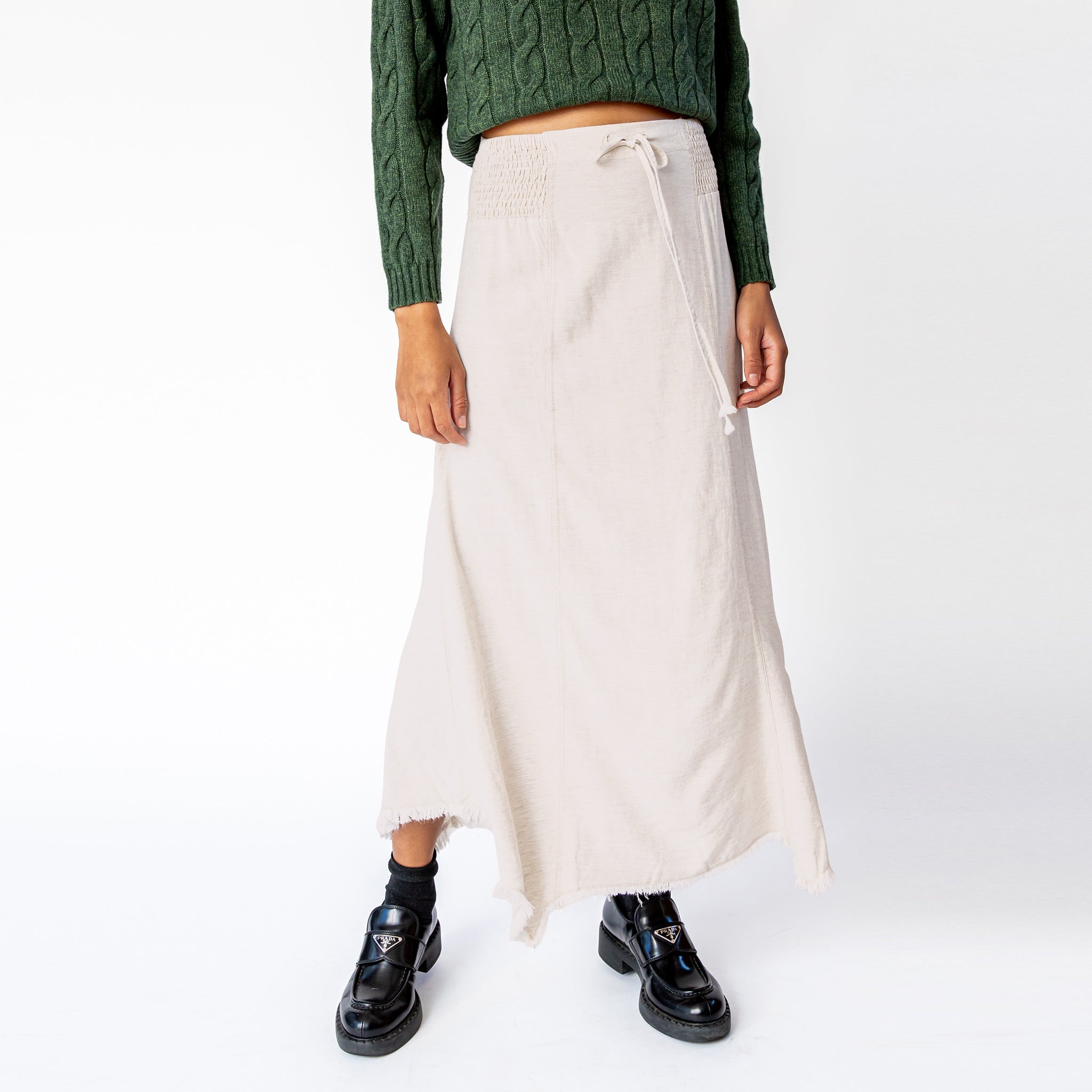A close up of a model wearing Misc Etc's smocked linen midi skirt in sand, featuring a smocked elastic waistband with drawstring and an asymmetrical hem, paired here with a green cable knit sweater and black loafers.