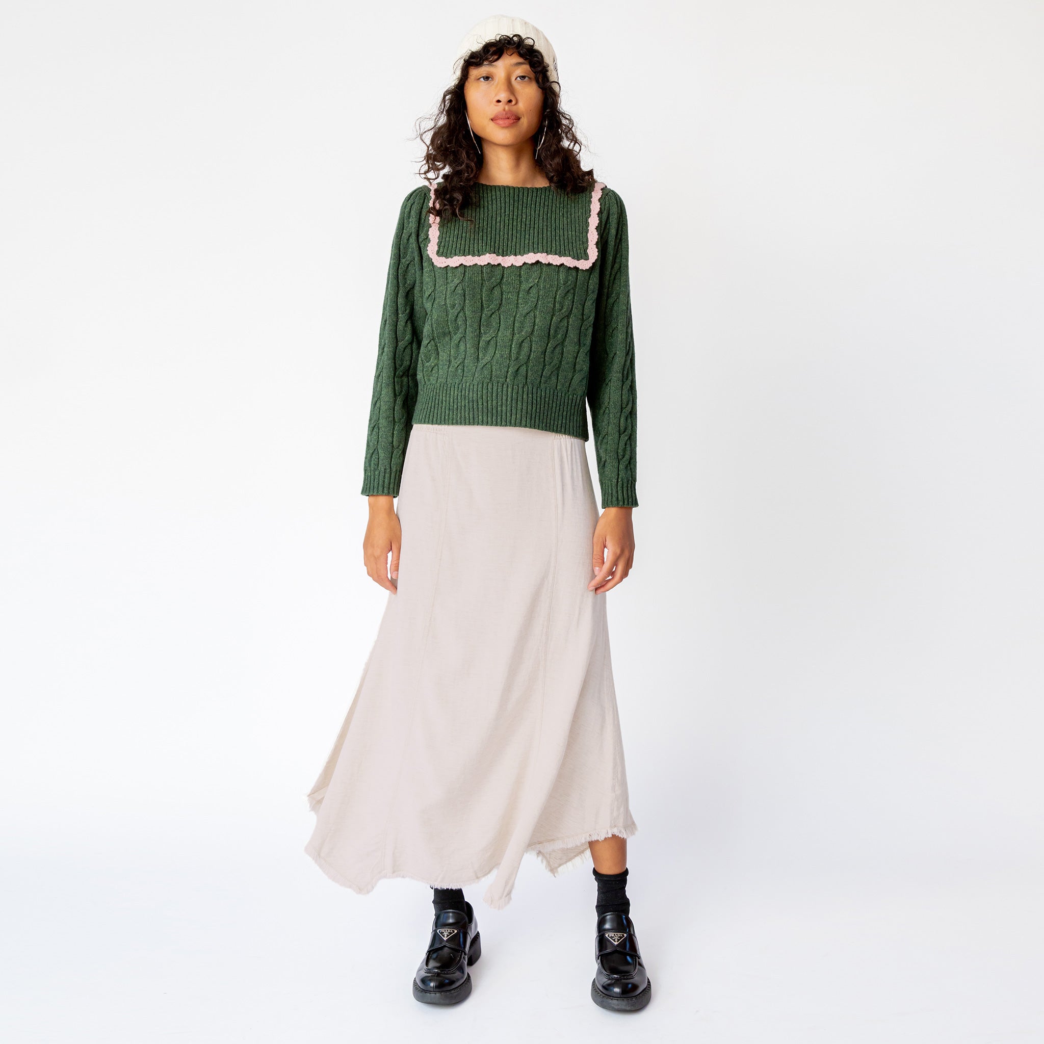 A model wears Misc Etc's smocked linen midi skirt in sand, featuring a smocked elastic waistband with drawstring and an asymmetrical hem, paired here with a green cable knit sweater and black loafers.