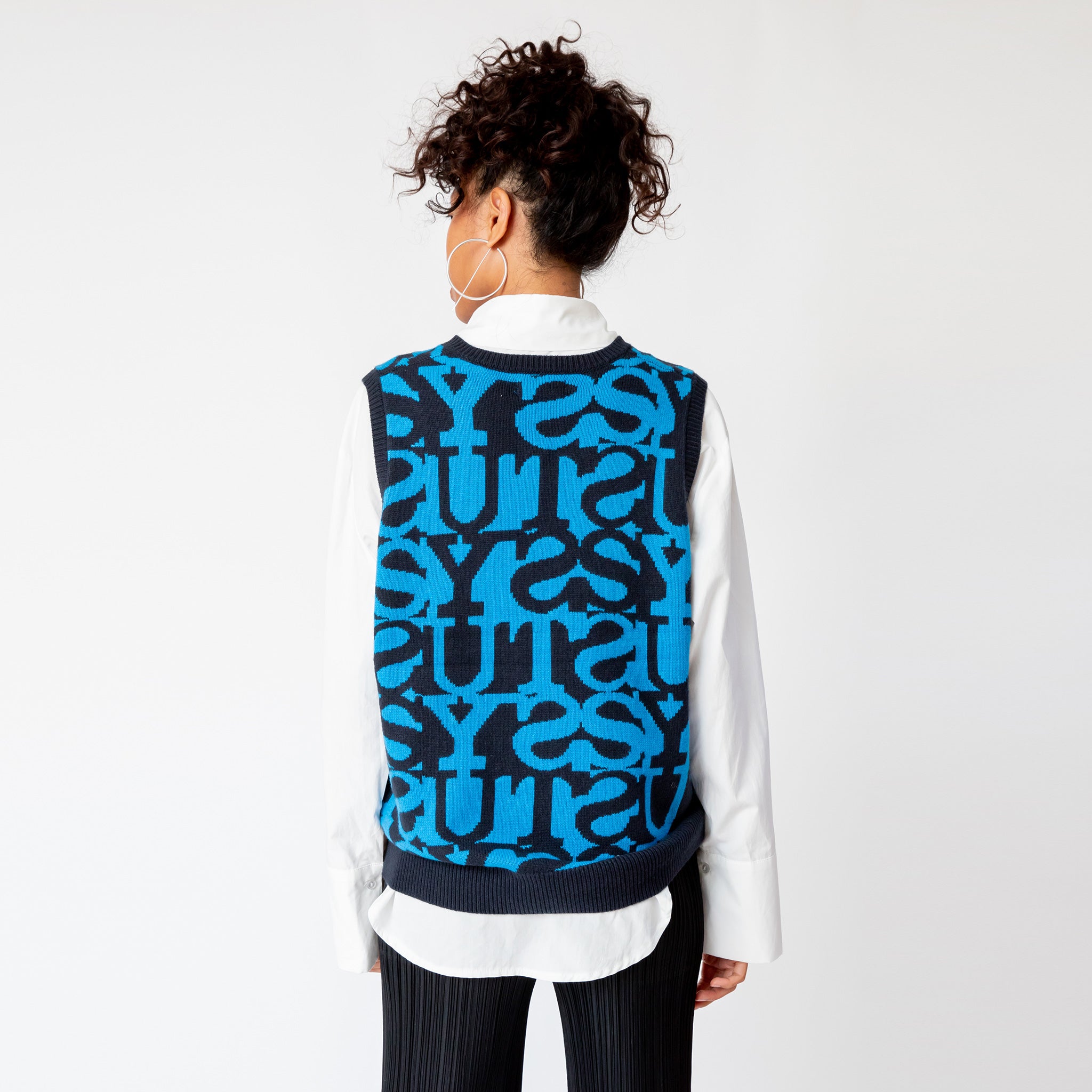 A model wearing the Stussy Stacked Sweater Vest - an oversized mens sweater vest with a blue and black allover repeating print of the word STUSSY - back view.