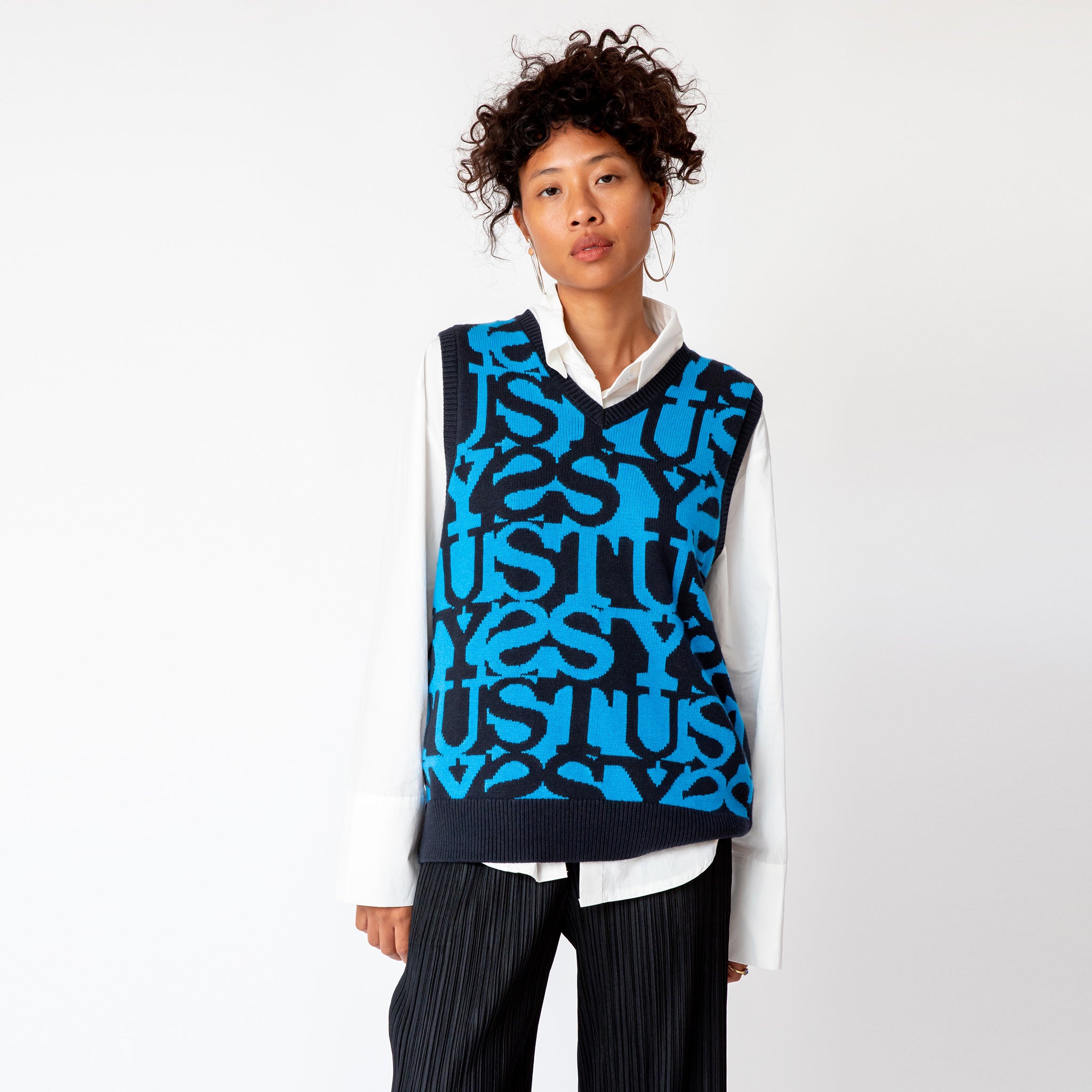 A model wearing the v-neck Stussy Stacked Sweater Vest - an oversized mens sweater vest with a blue and black allover repeating print of the word STUSSY.