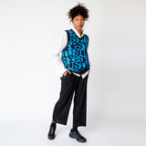 A model wearing the v-neck Stussy Stacked Sweater Vest - an oversized mens sweater vest with a blue and black allover repeating print of the word STUSSY, paired with pleated black trousers and black sneakers - full outfit view.