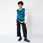 A model wearing the v-neck Stussy Stacked Sweater Vest - an oversized mens sweater vest with a blue and black allover repeating print of the word STUSSY, paired with pleated black trousers and black sneakers - full outfit view.