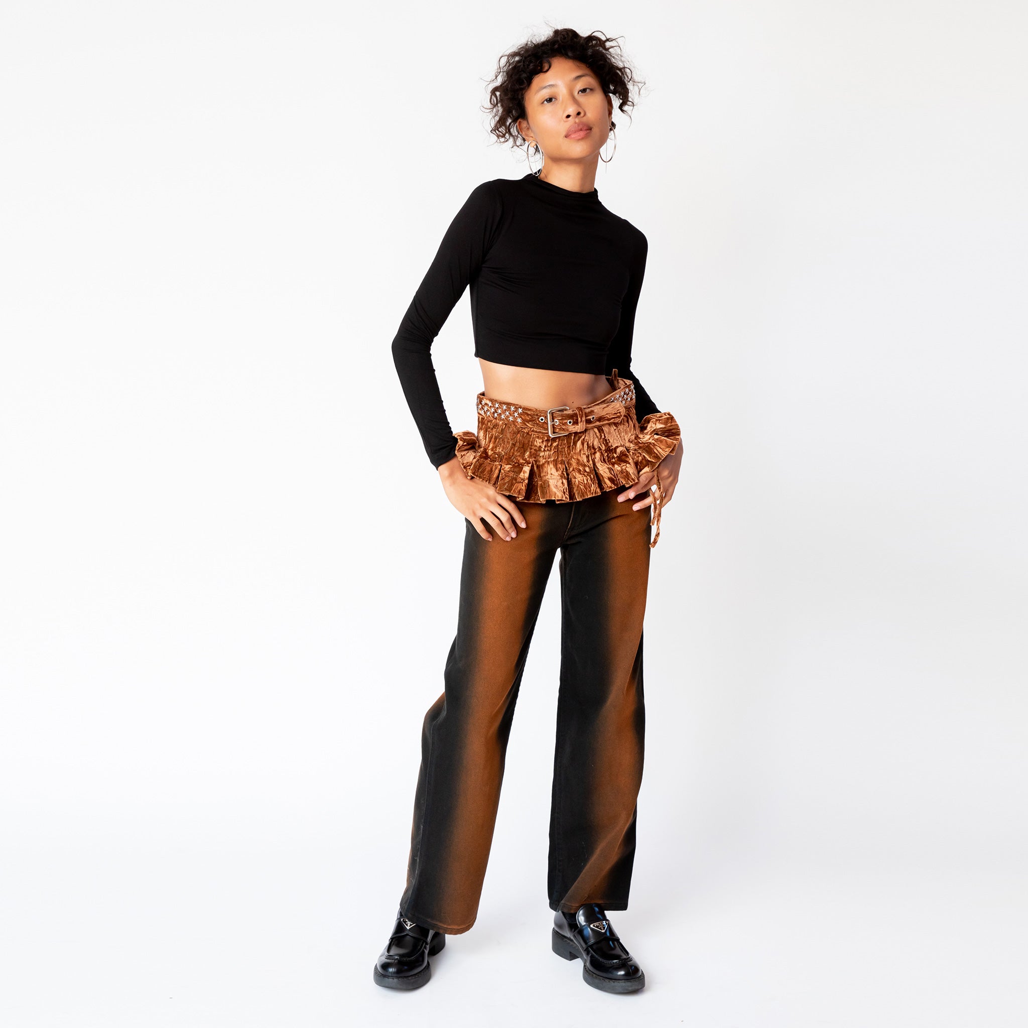 Full outfit view of a model wearing the velvet Star Pleated Belt in bark brown, an expressive belt with studded metal stars along the waistband and descending pleated micro skirt, paired with a longsleeve black crop top and gradient jeans.