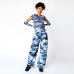 A model wears the blue and white graphic dolphin printed, wide leg Stomp Pant by Collina Strada with matching mesh top and gloves.