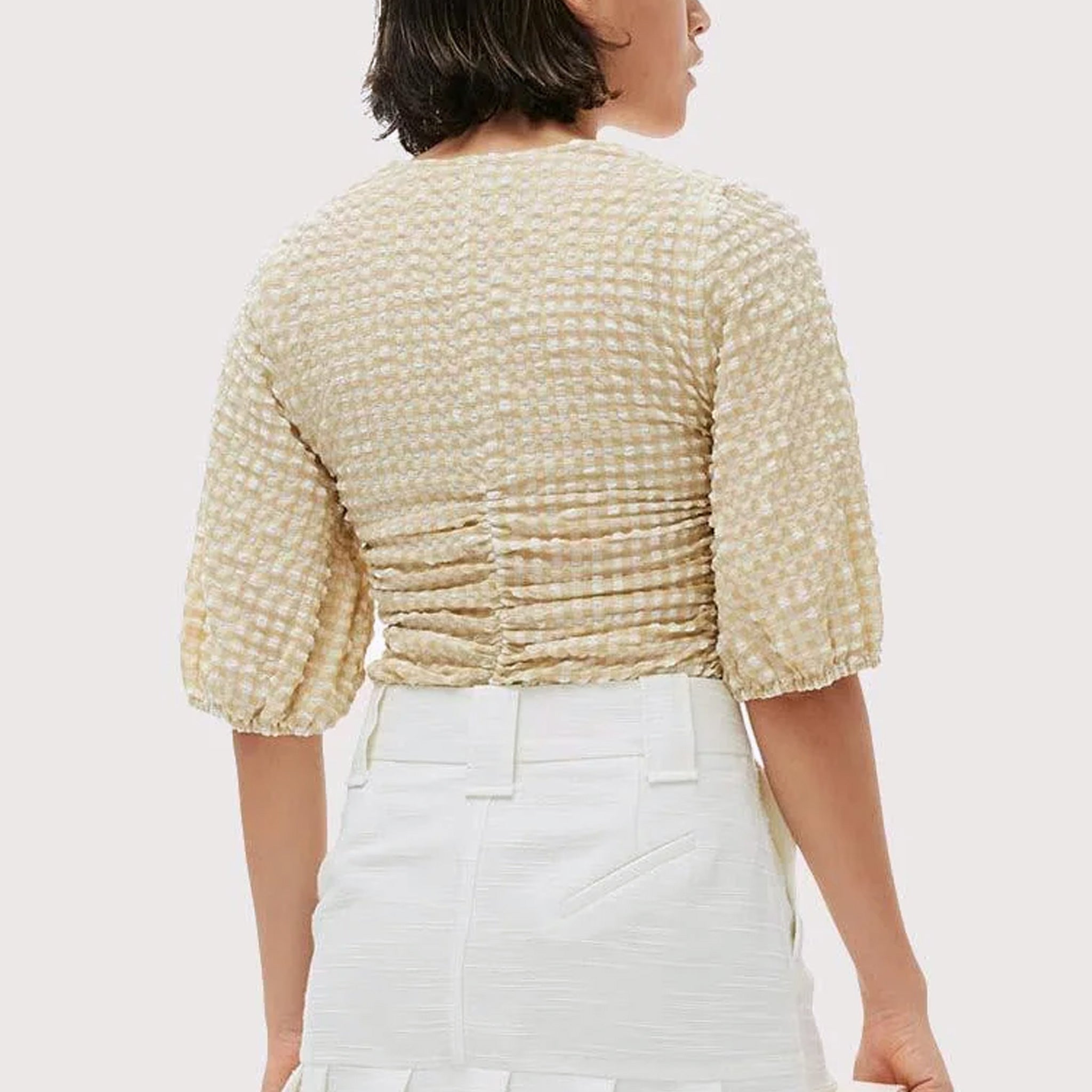 A model wears the beige and white seersucker gathered blouse by Ganni tucked into white bottoms - back view.