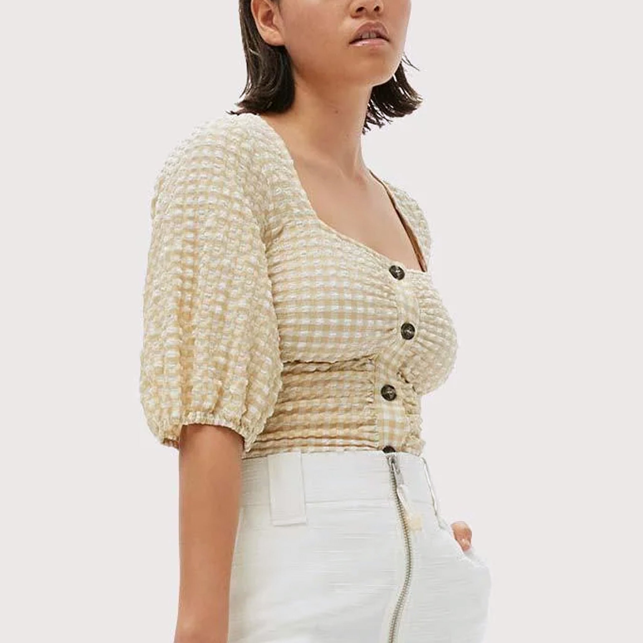 A model wears the beige and white seersucker gathered blouse by Ganni tucked into white bottoms.