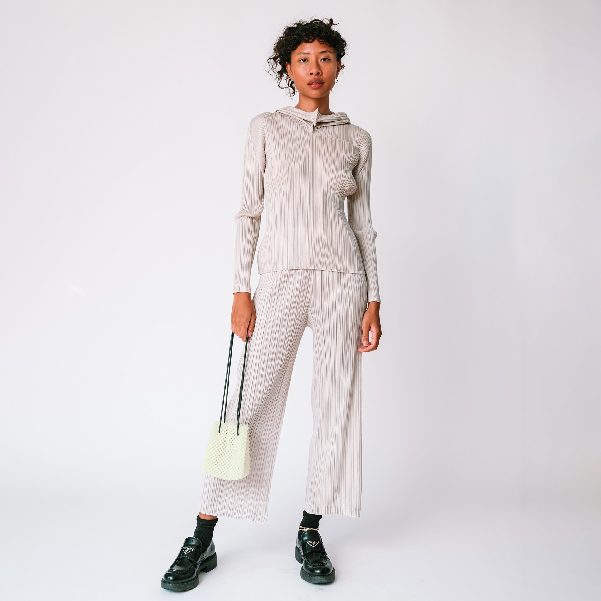 A model wears the light grey pleated Thicker Bottom 2 Pants by Pleats Please with matching hoodie and black loafers while carrying a beaded bag.