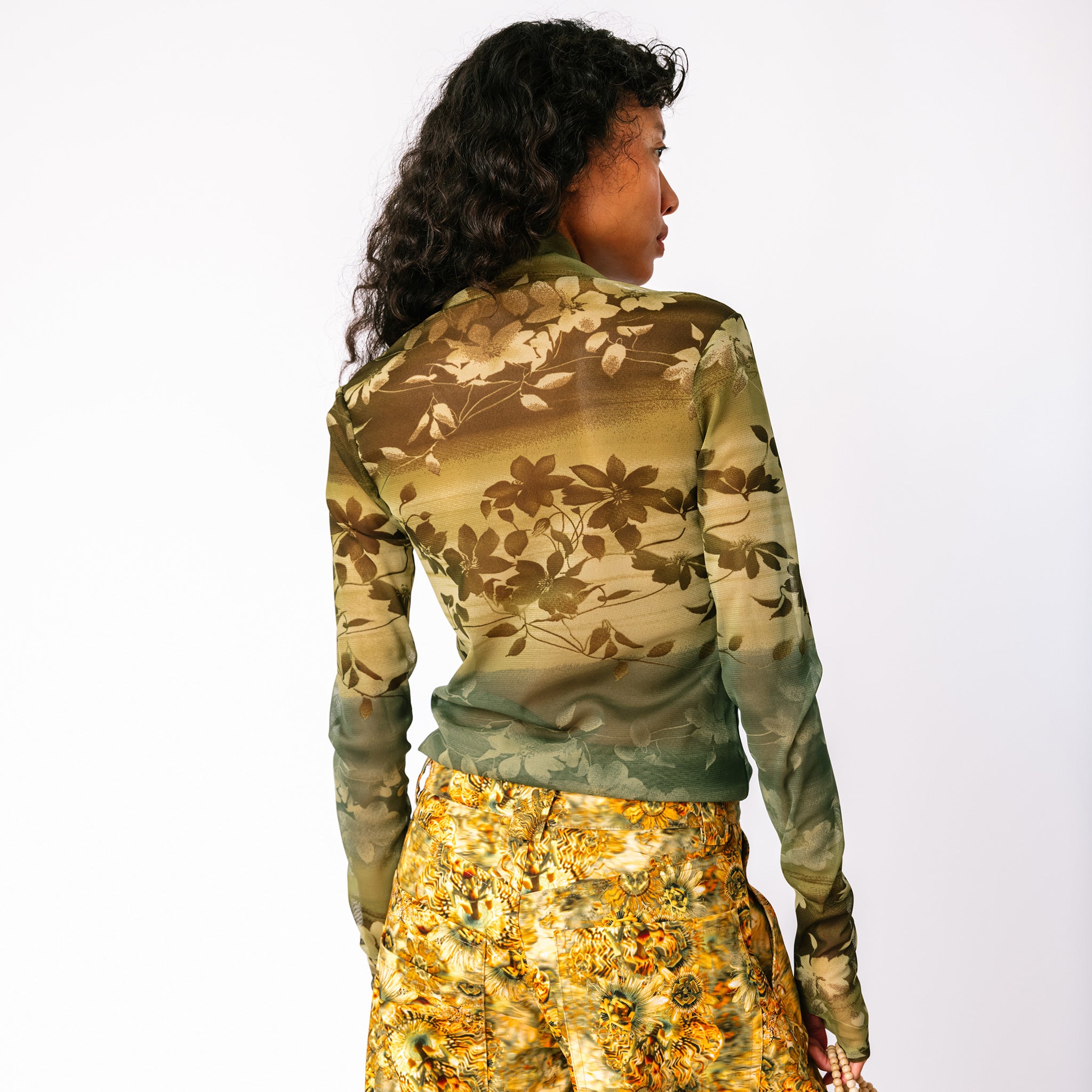 A model wears the long sleeved mesh Thumbtastic Top in an ombre green print with floral and leaves silhouettes - back view.