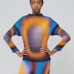 A lightweight long sleeve pleated top with a vibrant radial printed all-over graphic with orange, black and blue hues, worn by a model with her hands on her waist.