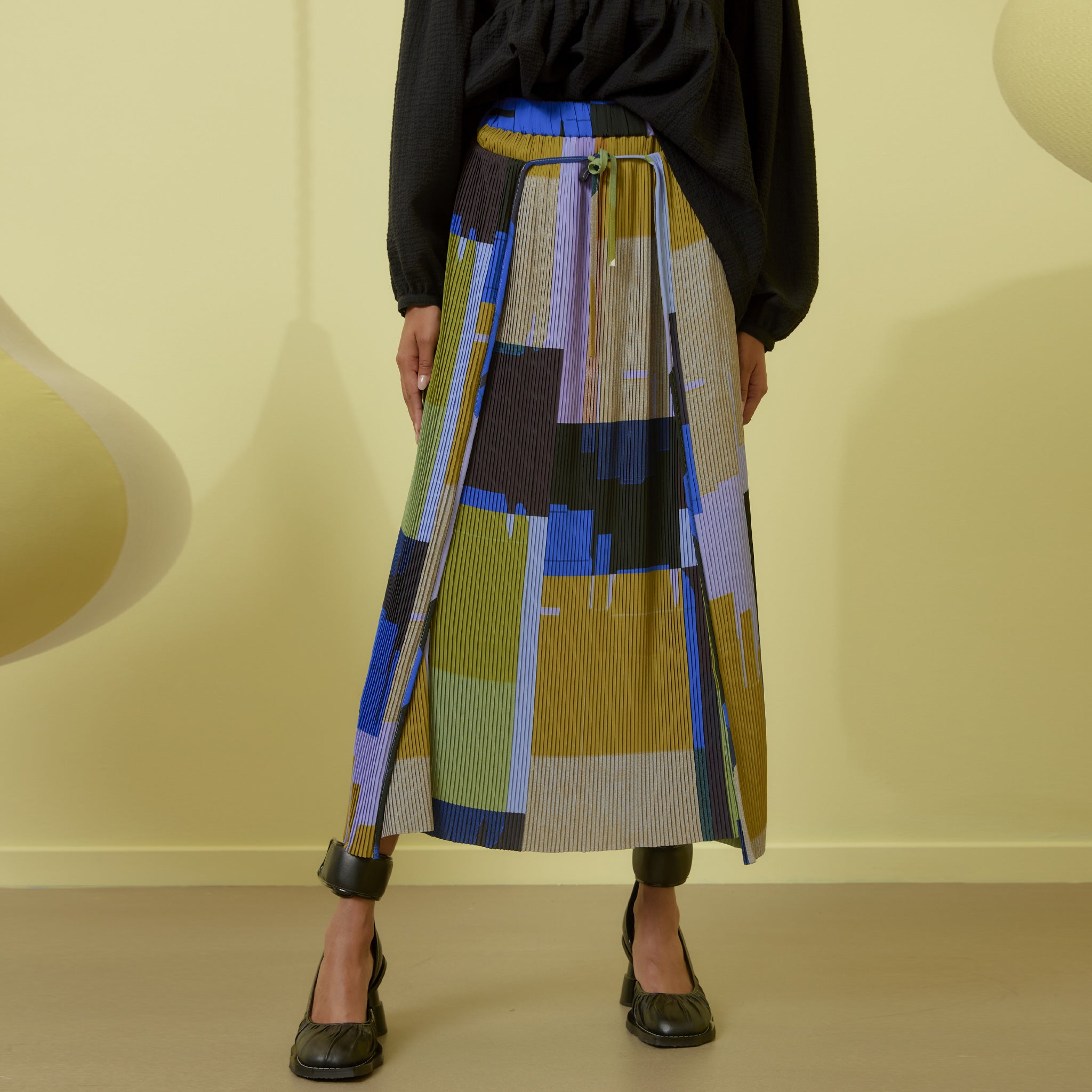 A pleated graphic printed wrap skirt with waist tie, featuring blue, black beige and green blocks.