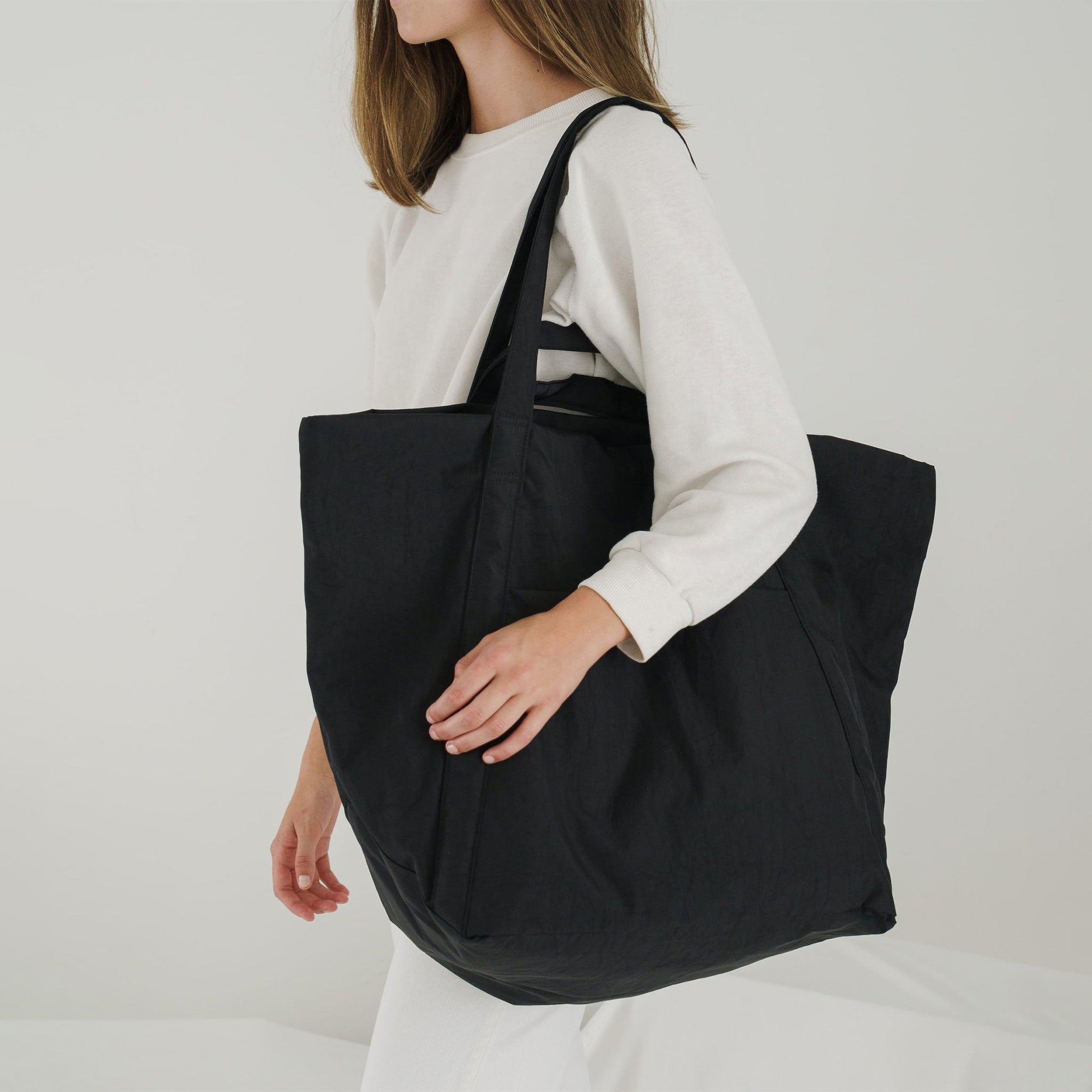 A large trapezoidal black nylon tote bag meant for traveling, fabricated in a puffy crinkly fabric, shown on the shoulder of a model.