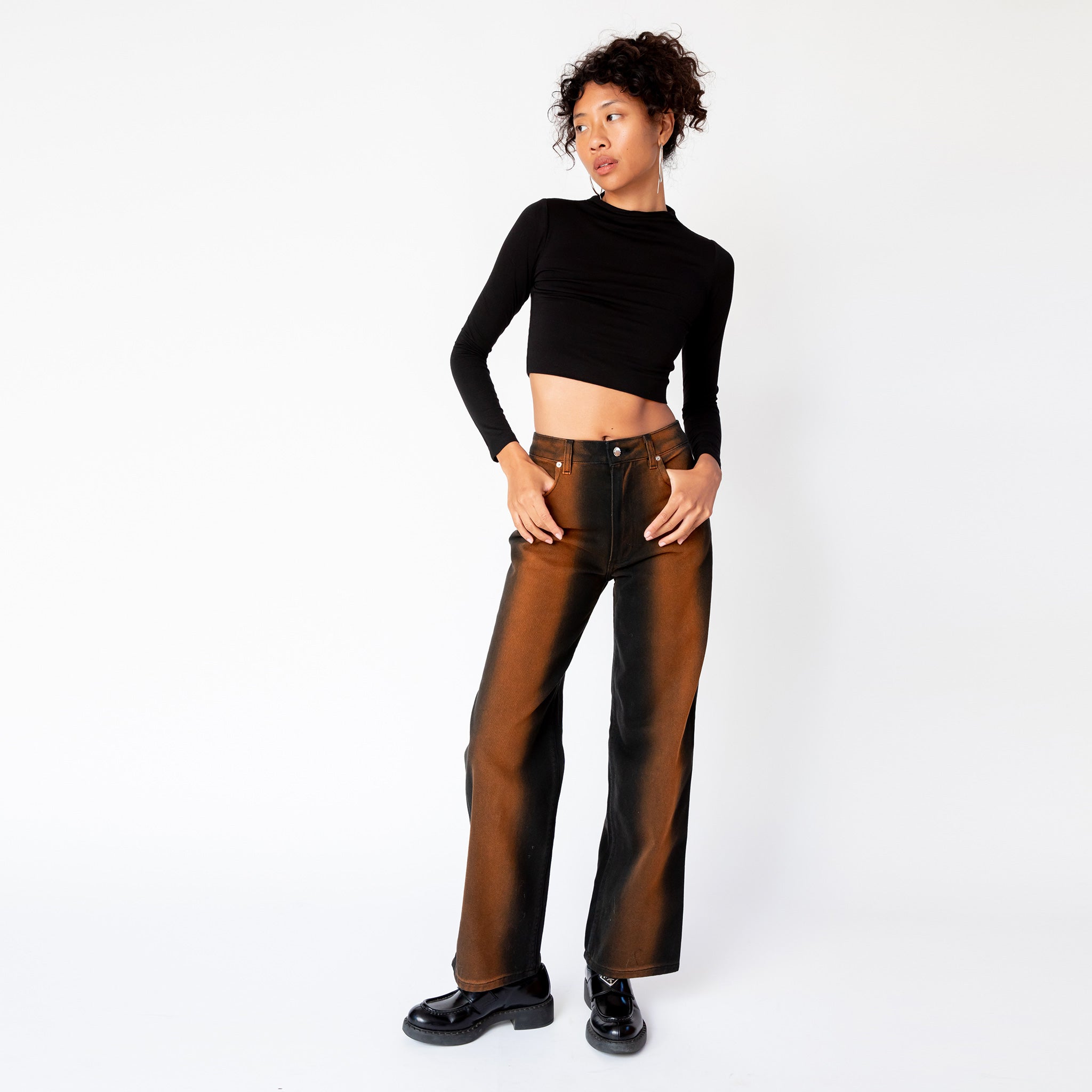 A model looks to the side while wearing Eckhaus Latta's wide leg jeans in ozone, featuring a vertical stripe of rust brown that gradients to black along the sides of the leg, paired with a cropped longsleeved black top.