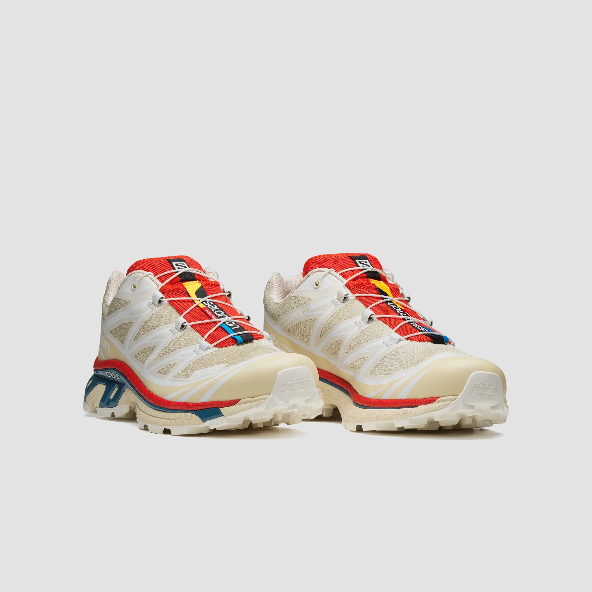 Off white XT-6 Salomon Sneakers with vibrant red and teal blue sole details, angled view of pair of shoes.