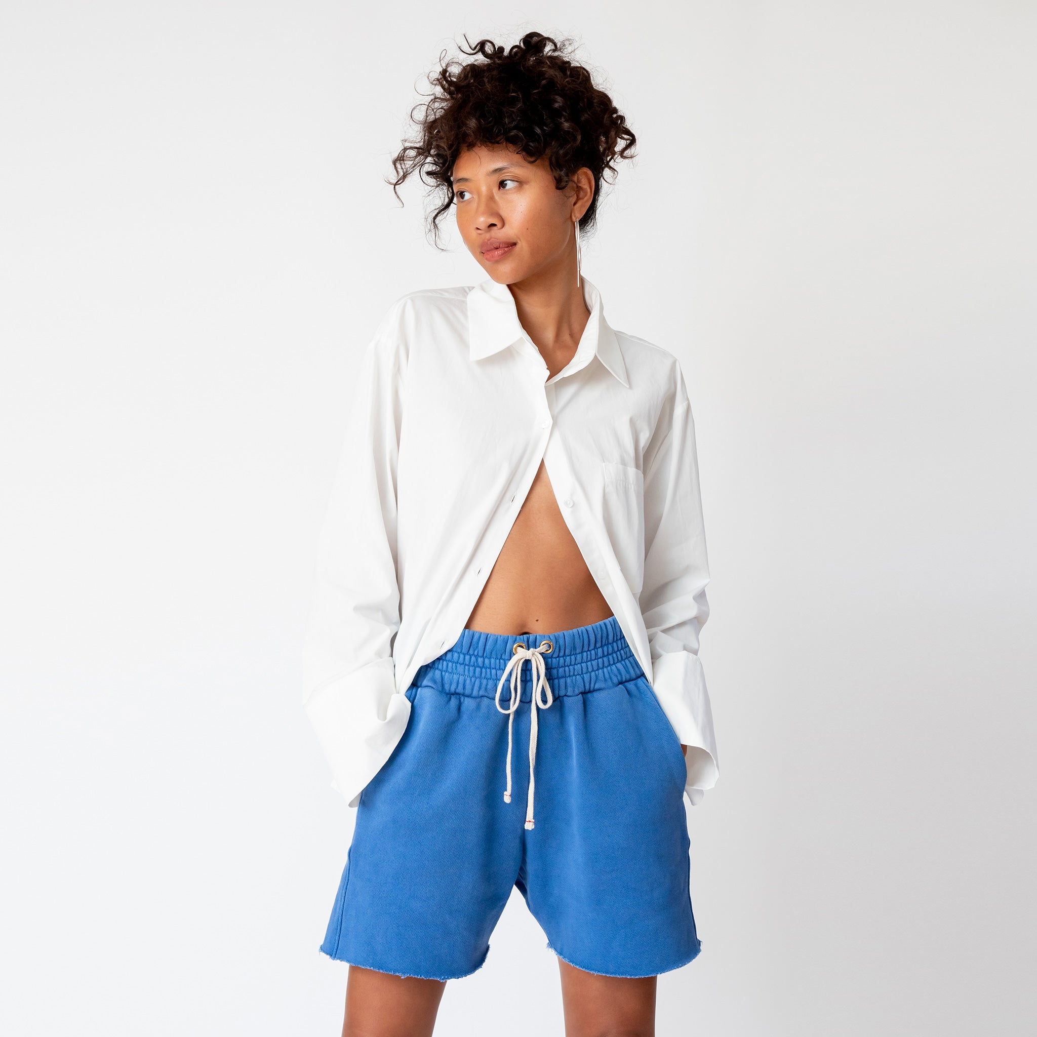 A model wears the Les Tien Yacht Short in electric blue, which feature a wide ruched elastic waistband, long white drawstrings, and an unfinished hem - paired here with a white button down shirt.
