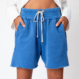 A model wears the Les Tien Yacht Short in electric blue, which feature a wide ruched elastic waistband, long white drawstrings, and an unfinished hem - front close up view.