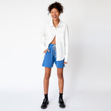 A model wears the Les Tien Yacht Short in electric blue, which feature a wide ruched elastic waistband, long white drawstrings, and an unfinished hem - paired here with a white button down shirt and black loafers.
