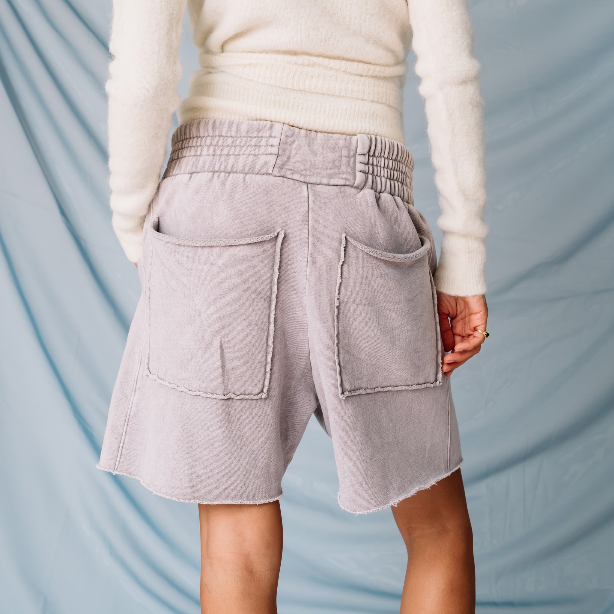 A model wears a white fuzzy sweater and the pale lavender stone Yacht Shorts by Les Tien against a soft blue backdrop - back view.