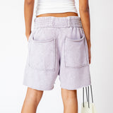 A model wears the pale lavender stone washed Yacht Shorts by Les Tien, featuring a wide elastic waist band, long white drawstrings and an unfinished raw hem - back view.