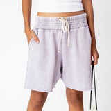 A model wears the pale lavender stone washed Yacht Shorts by Les Tien, featuring a wide elastic waist band, long white drawstrings and an unfinished raw hem - front view.