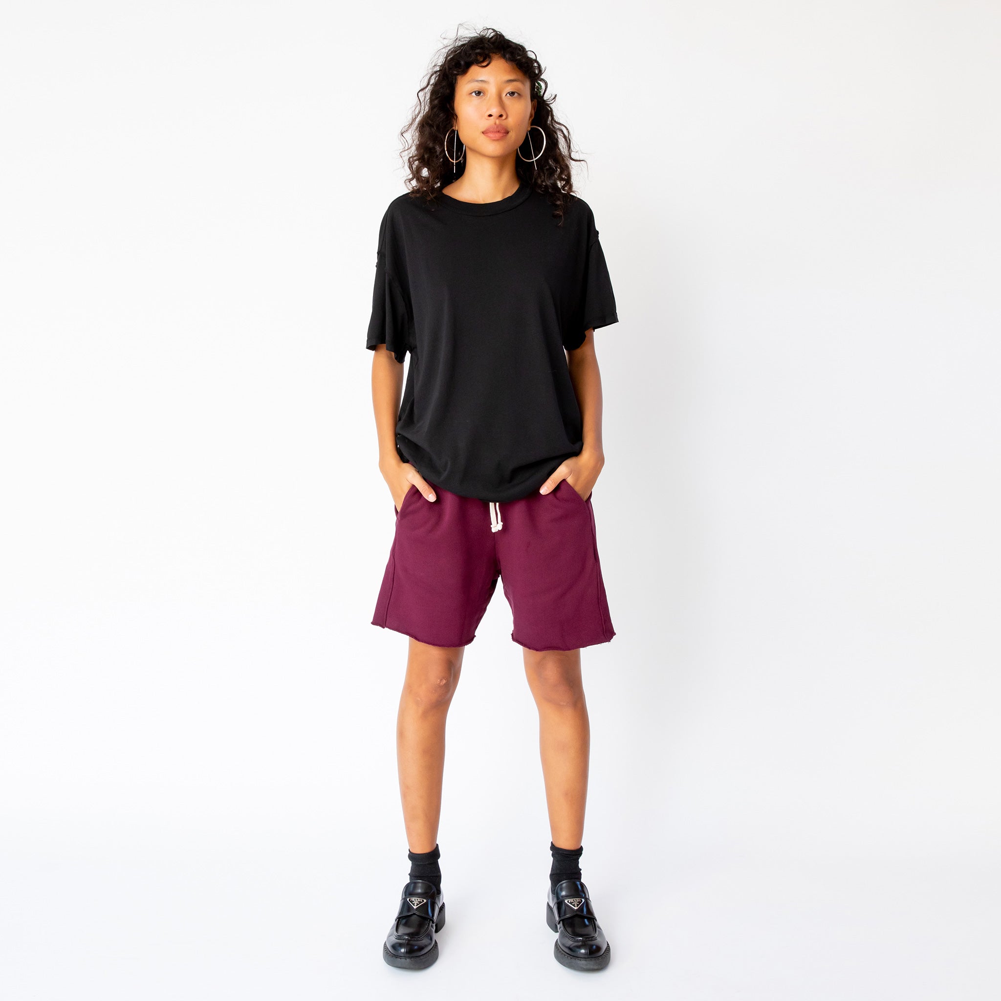 A model wears the merlot-red pigment dyed Yacht sweat shorts by Les Tien, paired with a black t-shirt and black loafers - full outfit view.