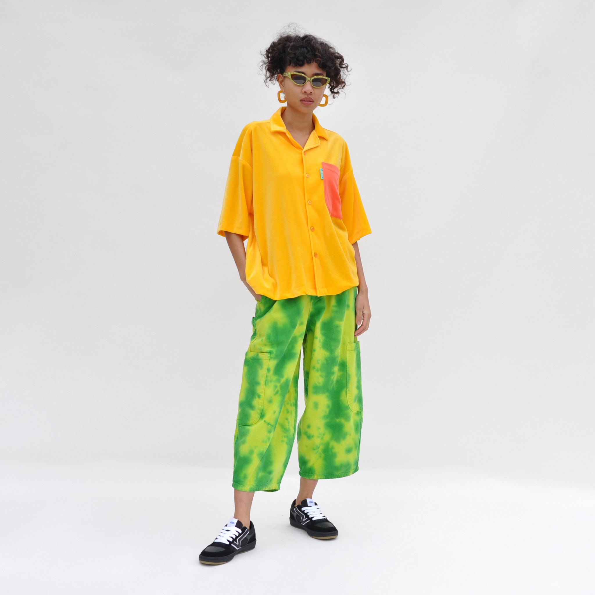 Full body image of a model wearing the chef pants by meals in watermelon. these oversized Cargo pants are green and yellow tie-dye. Like the outside of a watermelon.