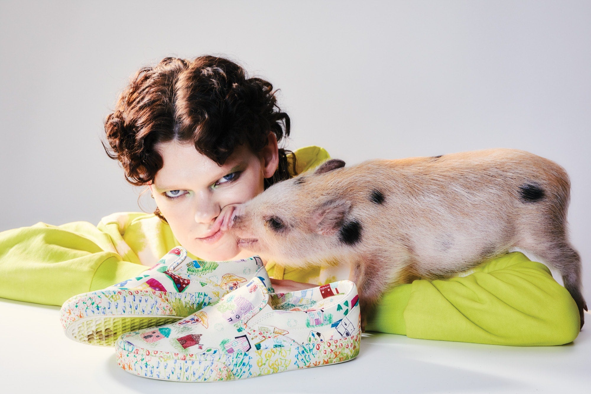 Close detail photo of a model laying next to a pig and a vans slip on shoe that is hand drawn.