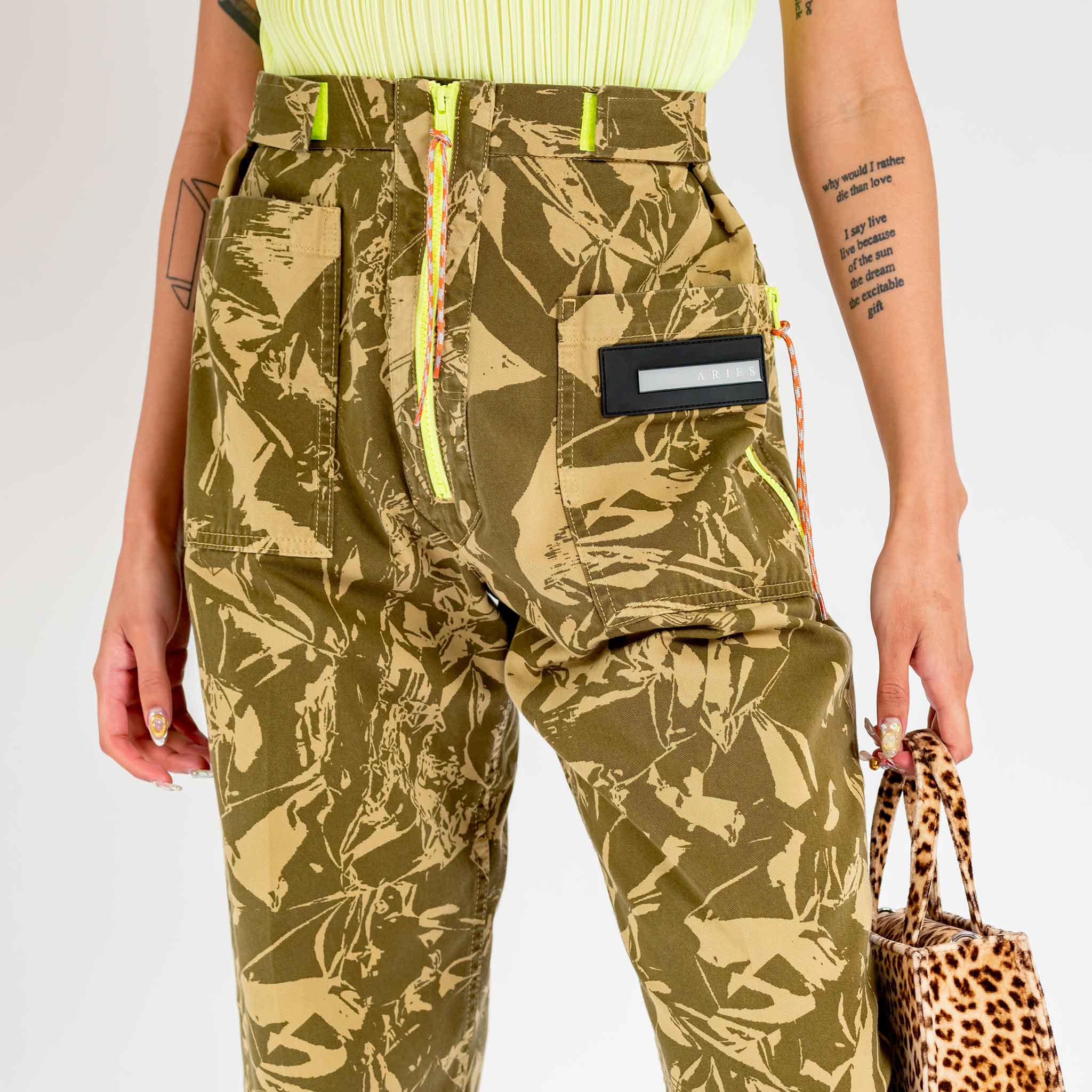Detailed close up photo of a model wearing the unisex Aries Crinkle Camo Walking Trouser, with fluorescent yellow zipper detail and belt loops.