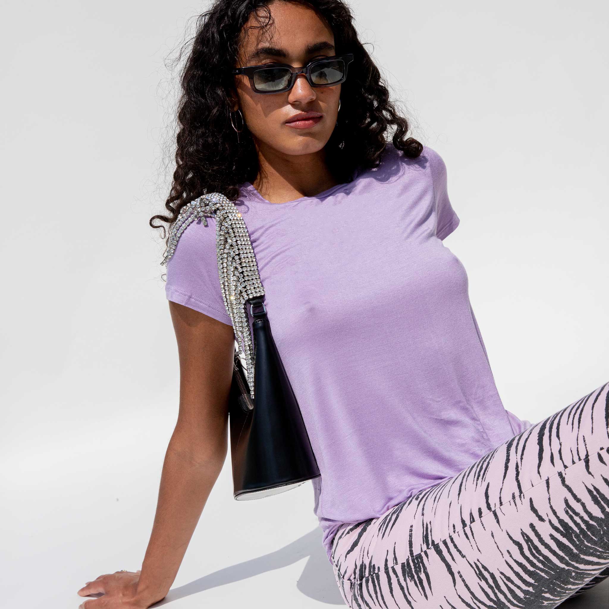 A model sits, leaning back with her hands while wearing the lavender Modal Tee, paired with zebra print pink jeans and a crystal-embellished shoulder bag.