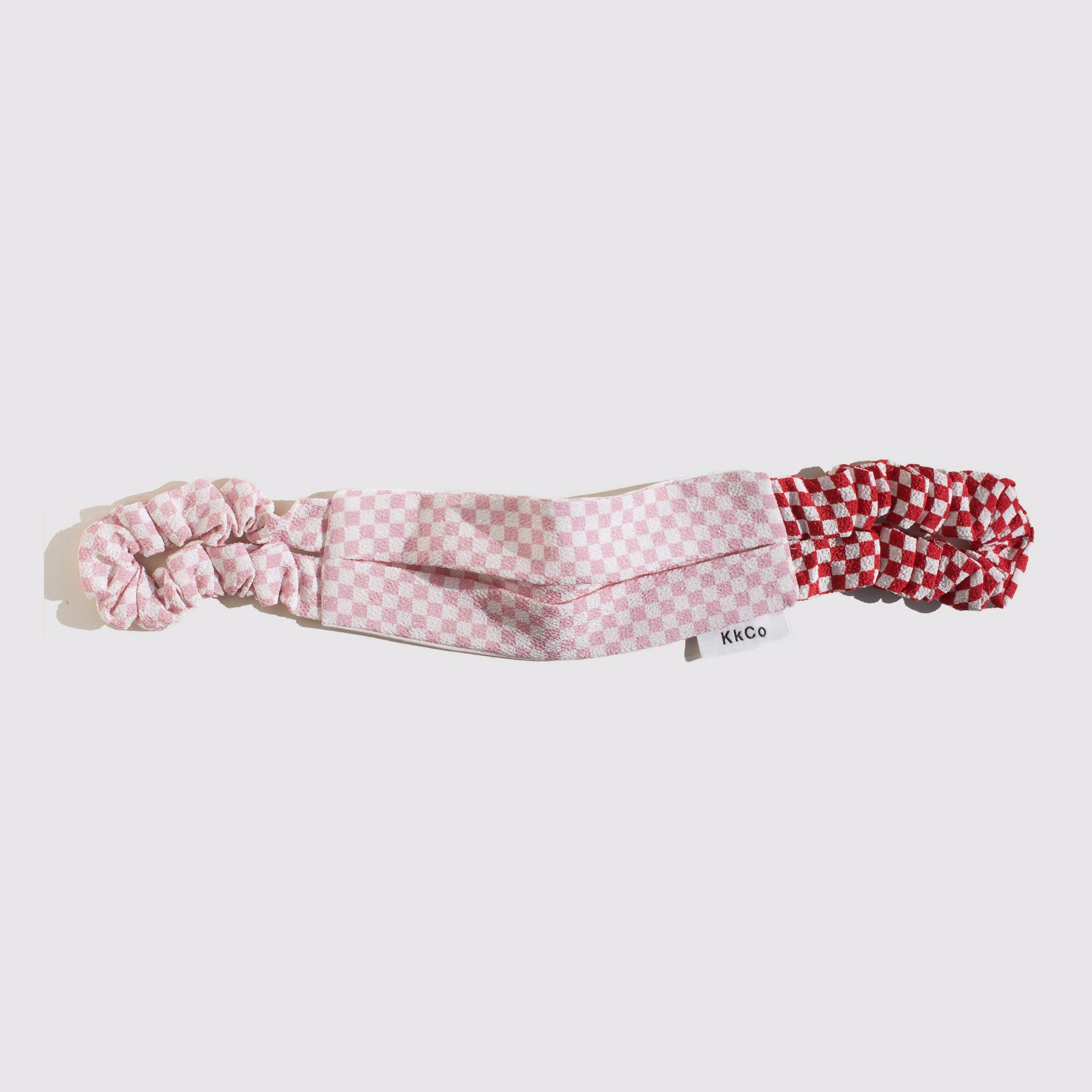 Flat photo of a pink and white checkered cloth face covering featuring scrunchie style ear loops, where the left ear loop's checks are red and white.