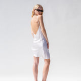 Model with her back to the camera looking over her shoulder as she wears a white mini slip dress.